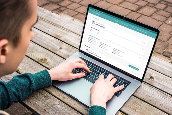 Ein braunhaariger Mann im grünen Pullover arbeitet an einem Mac im Freien an einem Holztisch sitzend. Auf dem Monitor ist die Software objego geöffnet.