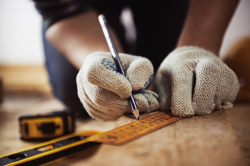 Nahaufnahme der Haende einer Person mit Arbeitshandschuhen, die mit einem Bleistift entlang eines Lineals auf ein Stueck Holz zeichnet. Links neben der Person liegen eine Wasserwaage und ein Maßband.