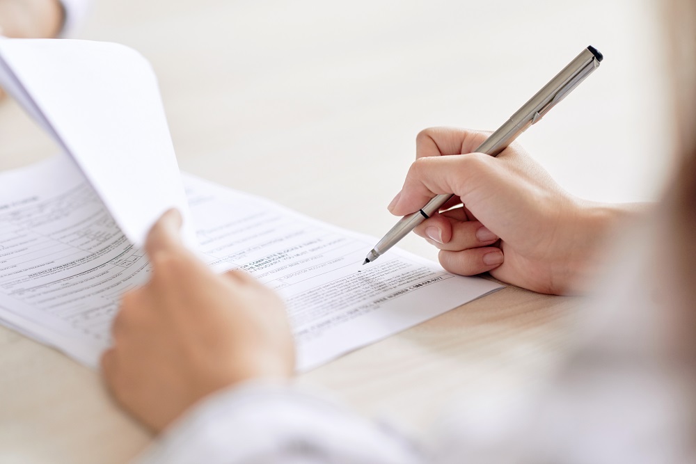 Haende einer Person, die mit der einen Hand ein Dokument bestehend aus mehreren Seiten hochhaelt und mit der anderen Hand einen Stift ansetzt.