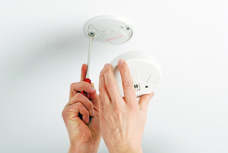 Haende bringen einen Rauchmelder an einer weißen Decke an. Die eine Hand dreht eine Schraube in die Decke durch den fest angebrachten Teil des Rauchmelders. Die andere Hand umfasst den Teil des Rauchmelders mit der Batterie.