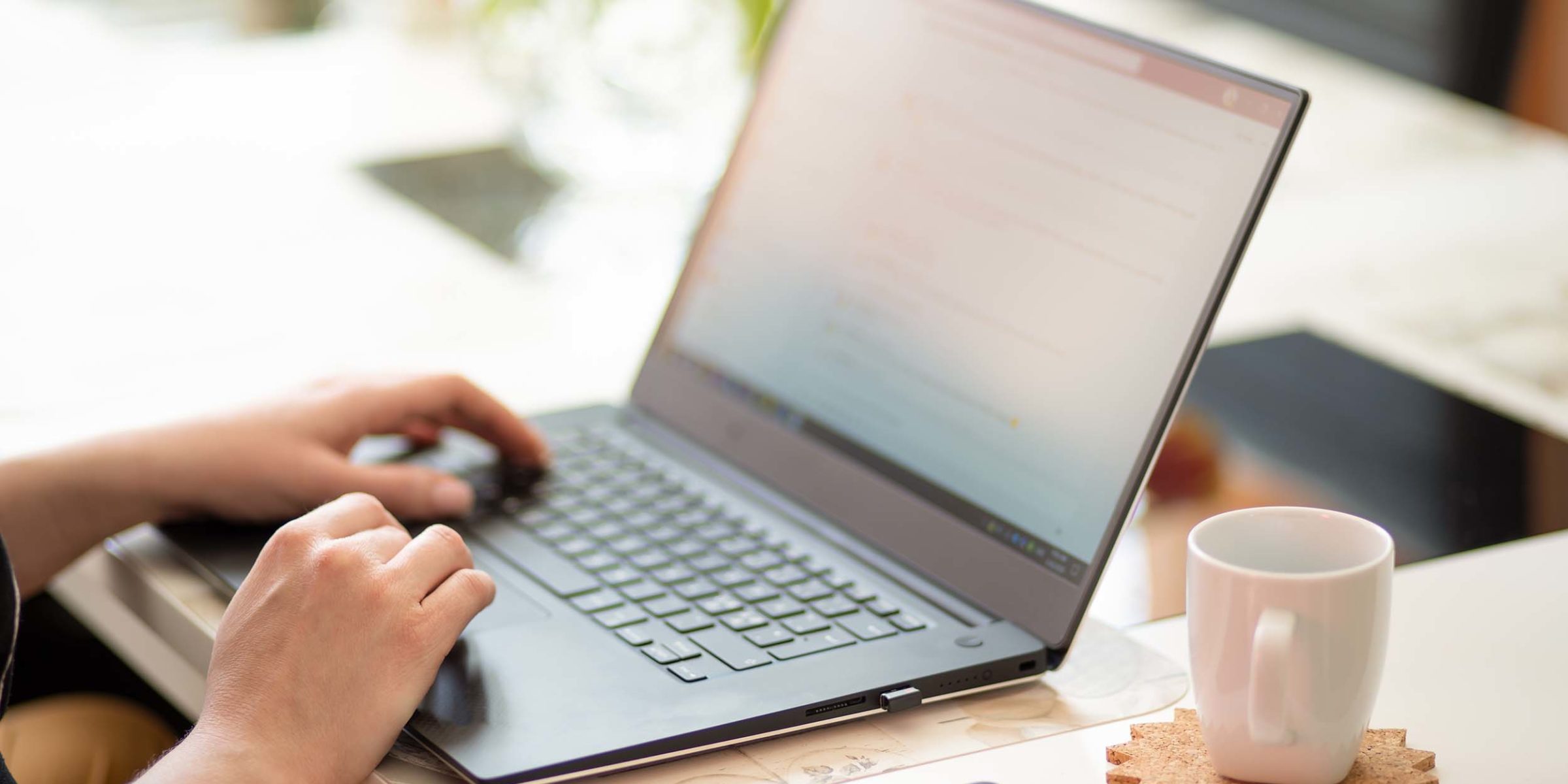 Haende einer Person, die an einem Schreibtisch sitzt. Die Haende liegen beide auf einem Laptop. Neben dem Laptop liegt ein Handy und eine weiße Tasse steht dort. Im Hintergrund ist eine Gruenpflanze zu erkennen.