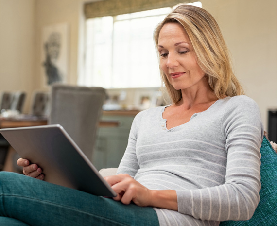 Frau sitzt auf einem Sofa und hat ein Tablet in der Hand. Mit einer Hand beruehrt sie den Bildschirm.