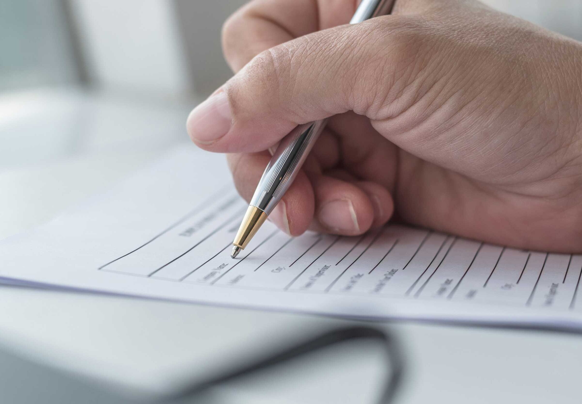 Hand, die einen Stift umfasst, setzt zum Schreiben auf einem Dokument an, das auf einem weißem Tisch liegt.