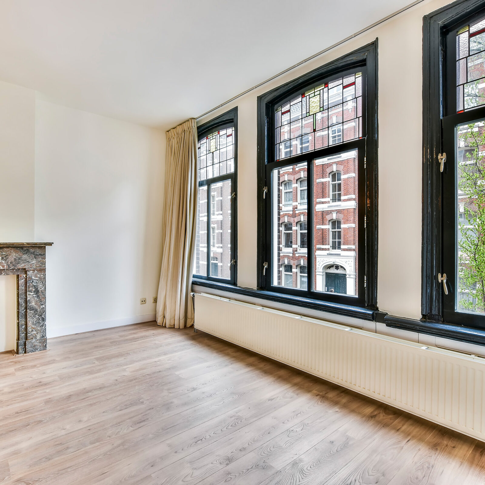Wohnungsleerstand: Schraeger Blick in ein leeres Zimmer einer Wohnung. Auf der rechten Seite ist eine grosse Fensterfront aus drei Fensterlementen und jeweiligem Oberlicht. Die Fenster haben dunkle Rahmen und die niedrige Fensterbank hat den selben Farbton. Darunter befindet sich ein langer, schmaler Heizkoerper, der fast ueber die gesamte Zimmerlaenge geht. An den Seiten sind beige Vorhaenge hoch angebracht, die ueber die gesamte Zimmerhoehe gehen. Die Waende sind weiss und der Boden aus hellem Holz mit Maserung. Auf der linken Bildseite ist ein Kaminsims, aber ohne Kamin.