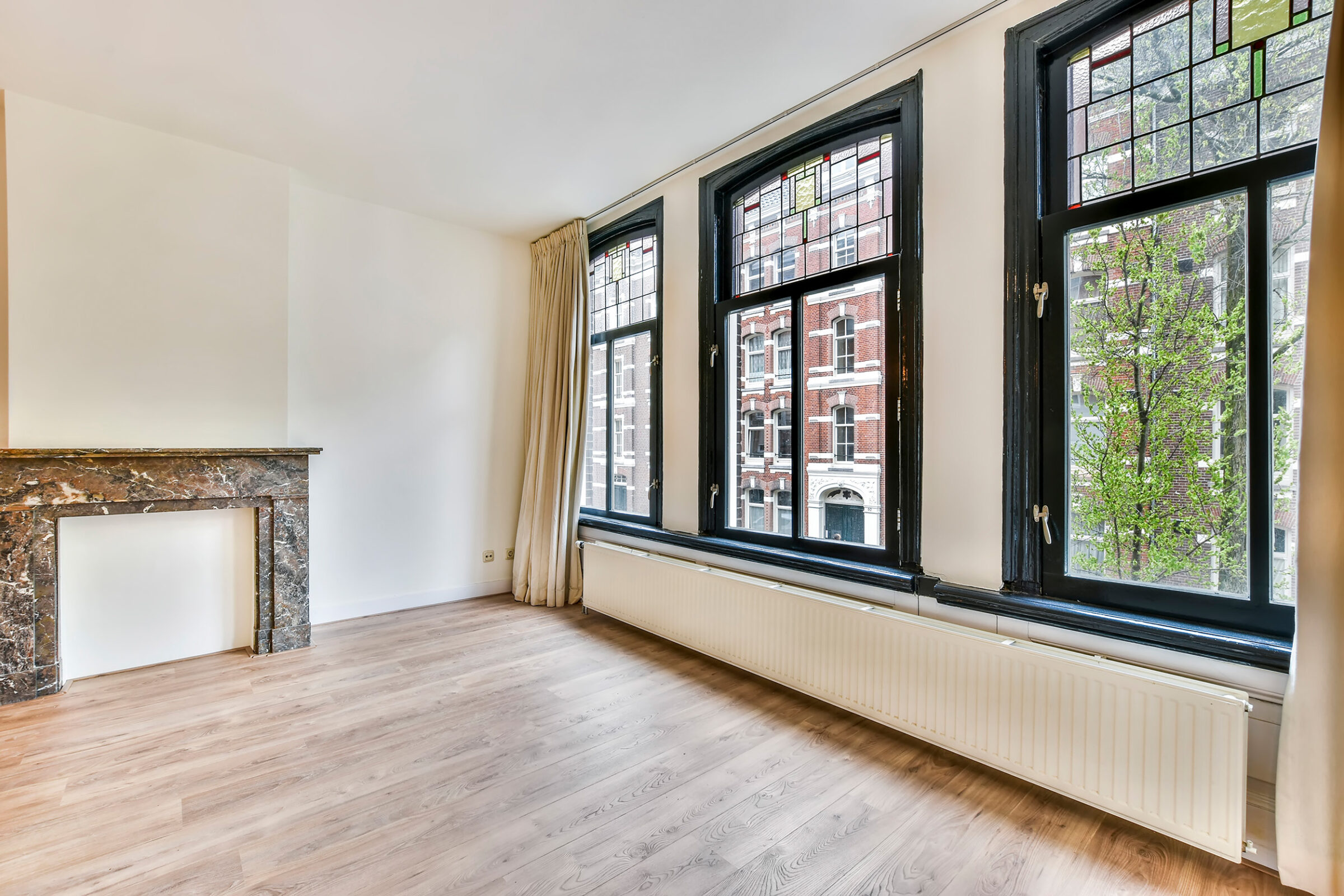Wohnungsleerstand: Schraeger Blick in ein leeres Zimmer einer Wohnung. Auf der rechten Seite ist eine grosse Fensterfront aus drei Fensterlementen und jeweiligem Oberlicht. Die Fenster haben dunkle Rahmen und die niedrige Fensterbank hat den selben Farbton. Darunter befindet sich ein langer, schmaler Heizkoerper, der fast ueber die gesamte Zimmerlaenge geht. An den Seiten sind beige Vorhaenge hoch angebracht, die ueber die gesamte Zimmerhoehe gehen. Die Waende sind weiss und der Boden aus hellem Holz mit Maserung. Auf der linken Bildseite ist ein Kaminsims, aber ohne Kamin.